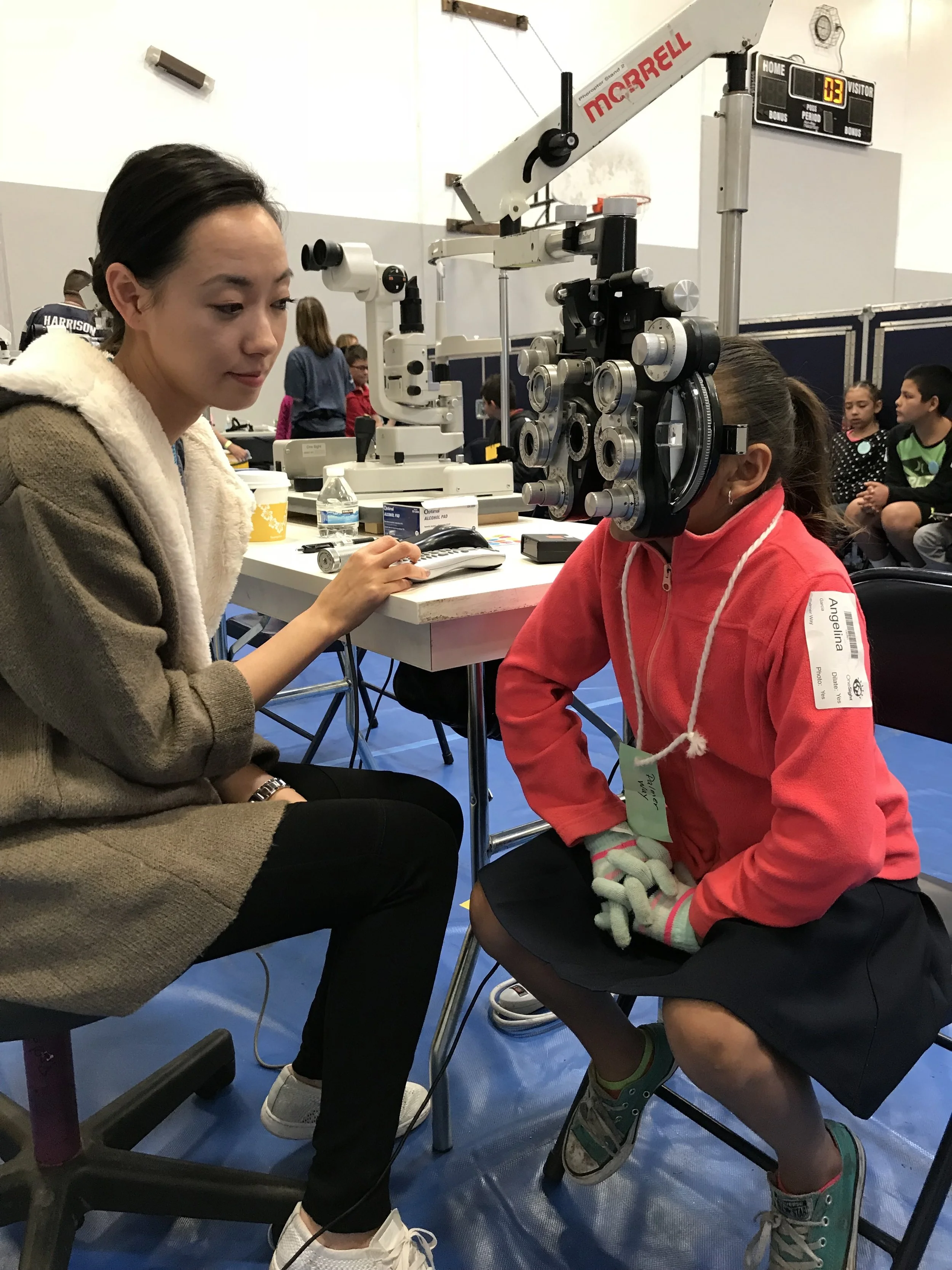 A New Vision Optometry doctor seated beside a young girl looking through a phoropter at a school vision screening in a gymnasium