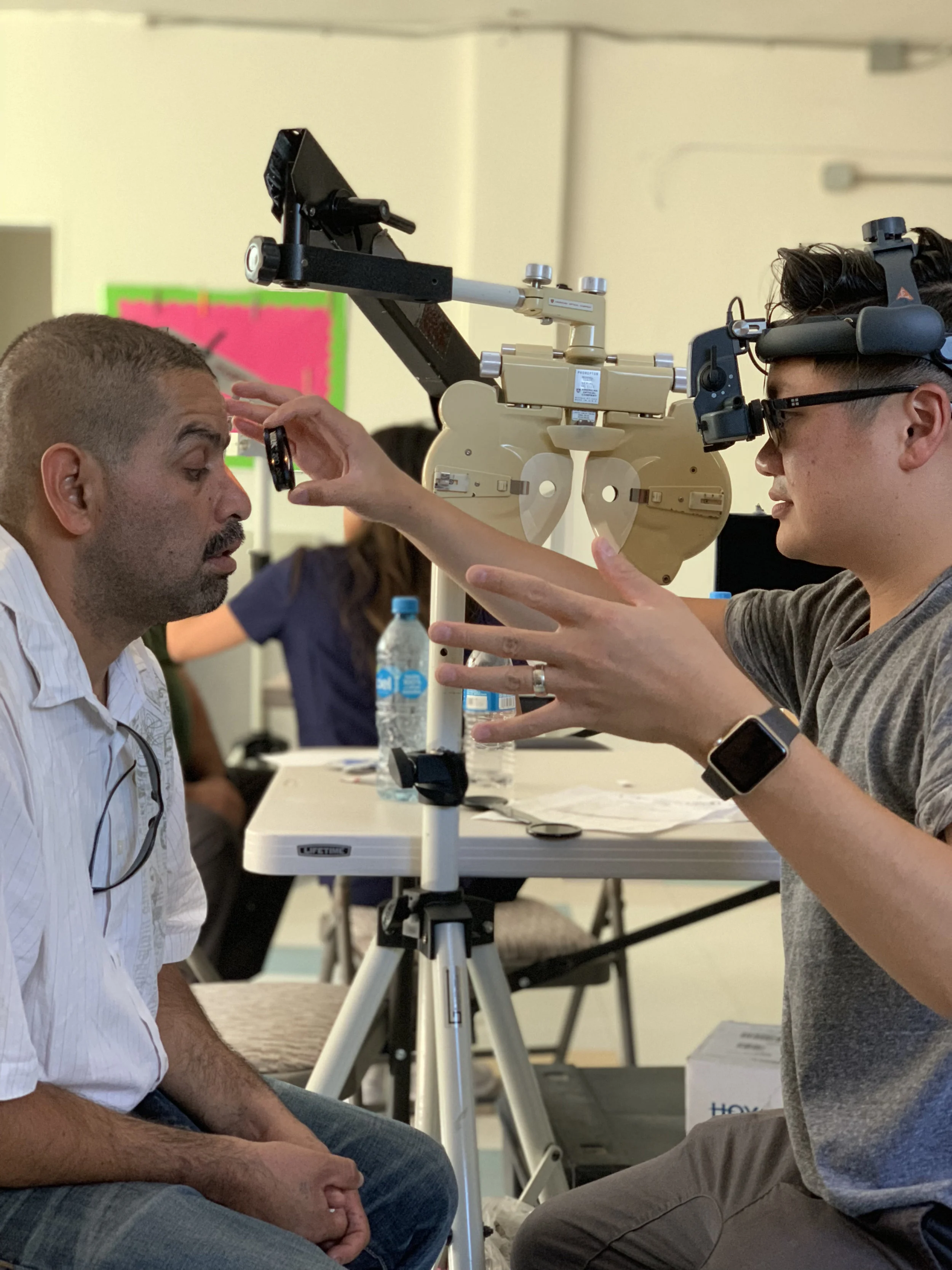 An optometrist wearing a head mounted examination light holds a trial lens in front of a patient's eye at the mission clinic, a portable phoropter on a stand between them
