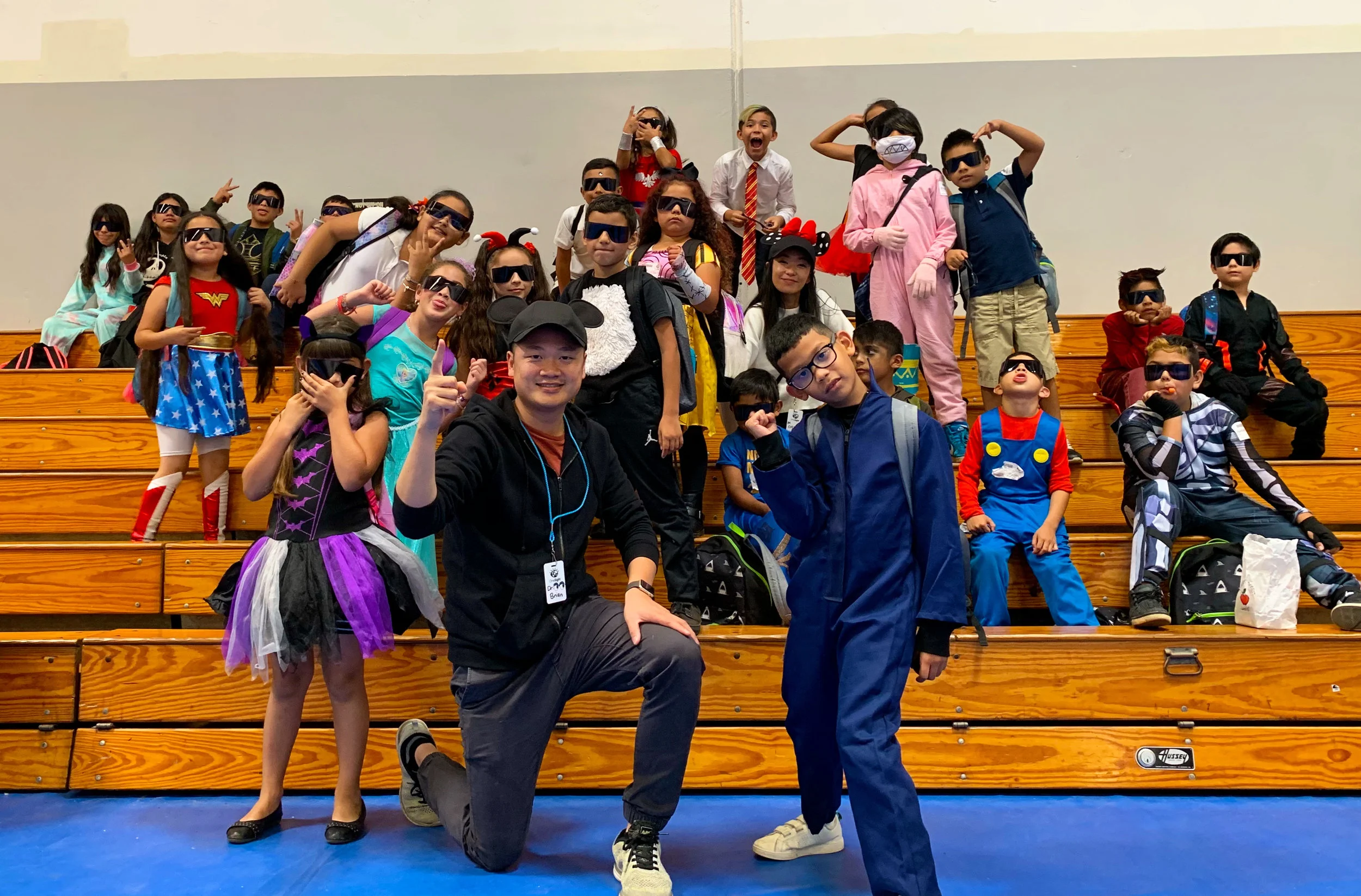 Dr. Nguyen kneeling in front of a school gymnasium full of children sitting on bleachers, the children in costume and wearing the dark shades handed out at a vision screening