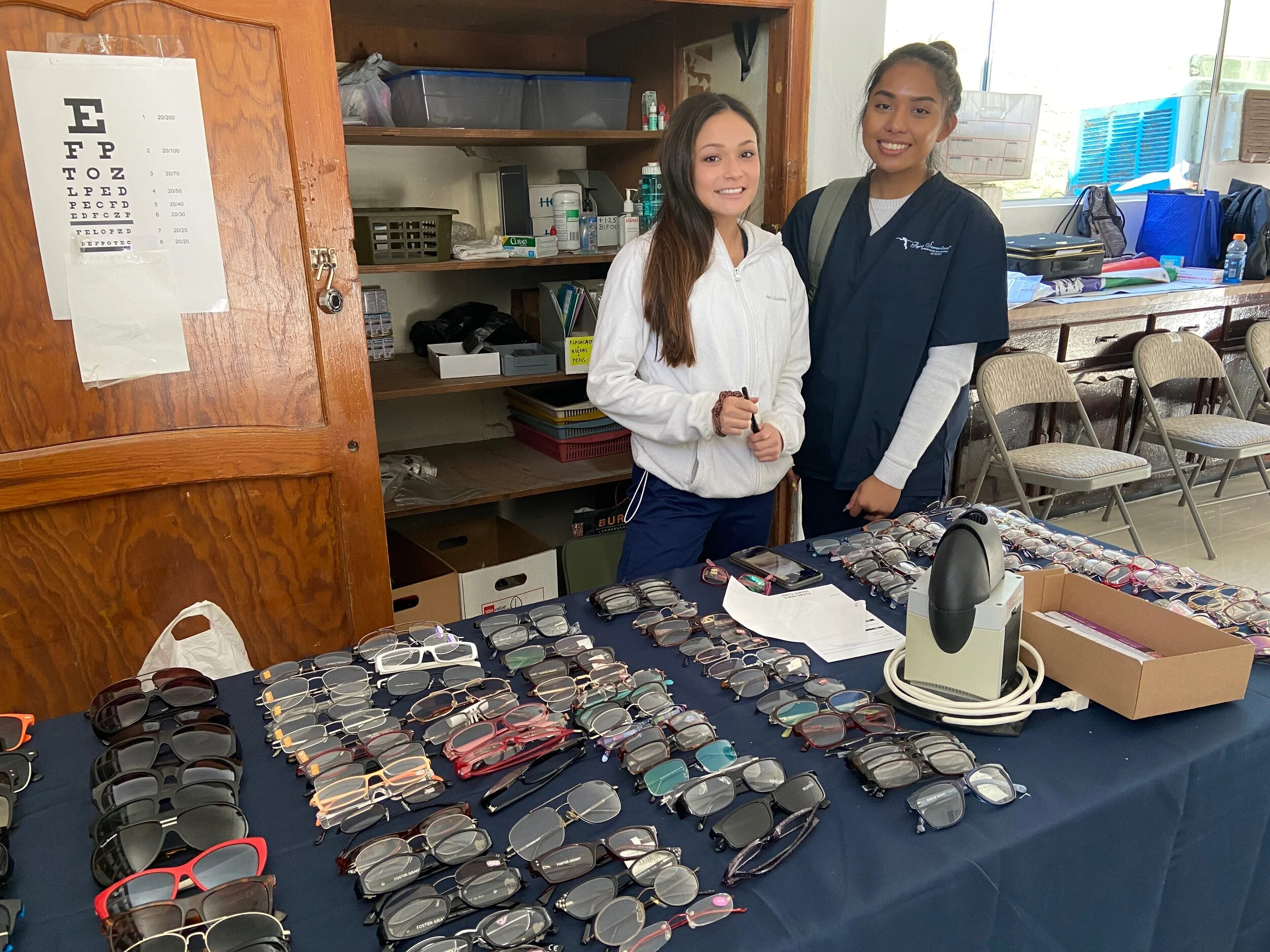 Two volunteers behind a table of eyeglass frames at the Tecate clinic optical department