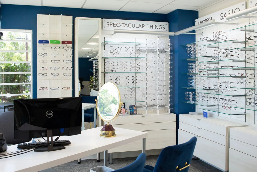 The dispensing desk at New Vision Optometry, a monitor and a brass framed mirror on a white desk in front of the frame displays and the Spec-tacular Things sign