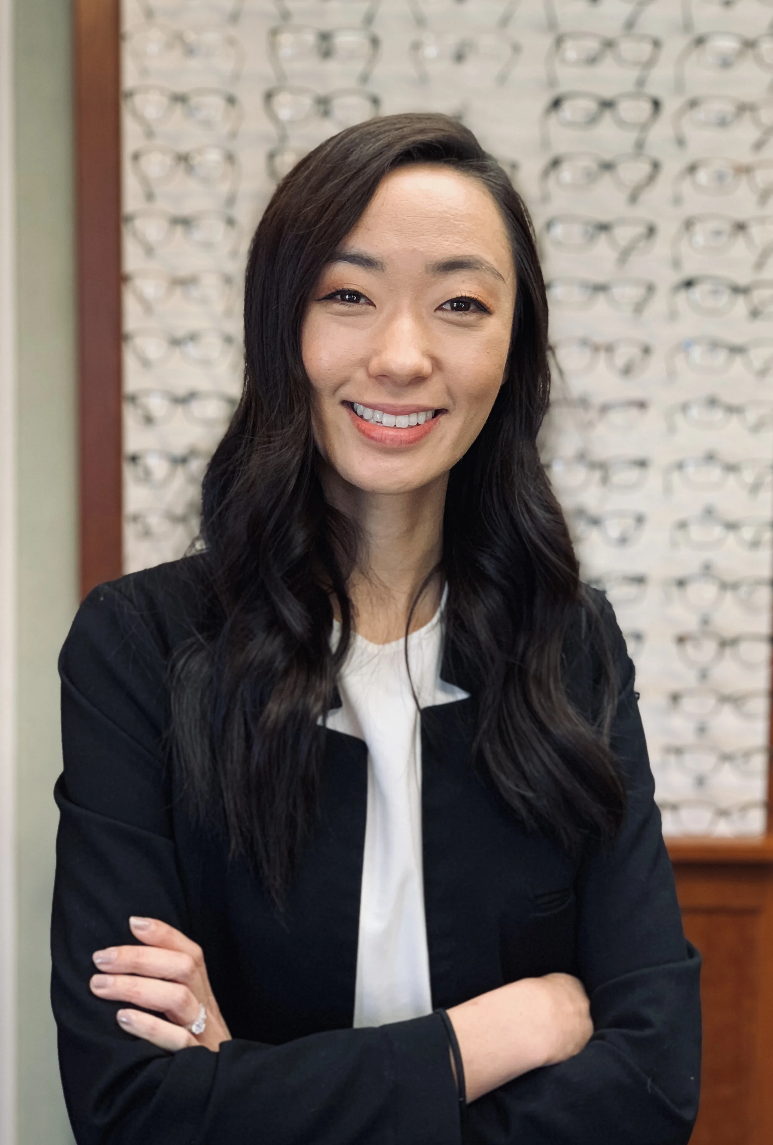 Dr. Eurri Choi in a black jacket, arms folded, standing in front of a wall of frames in the practice optical