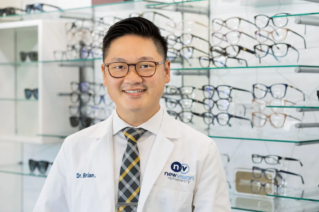 Dr. Long Nguyen, O.D., in a white coat with the New Vision Optometry logo, standing in the optical in front of the frame displays
