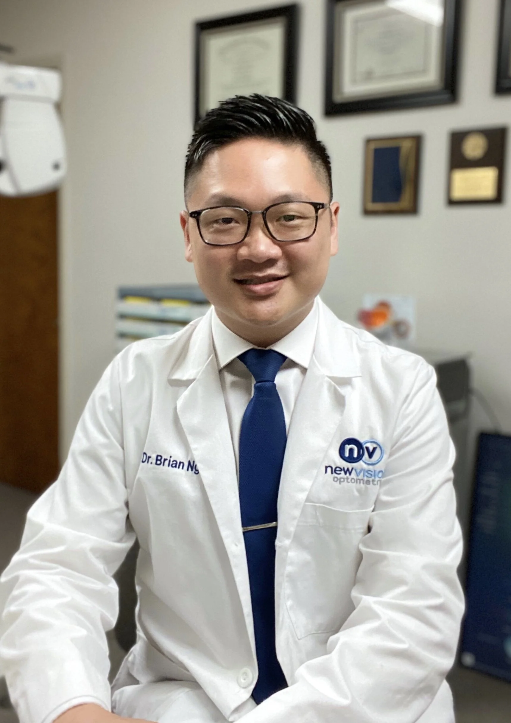 Portrait of Dr. Long Nguyen, O.D., wearing a white coat with the New Vision Optometry logo, framed diplomas on the wall behind him