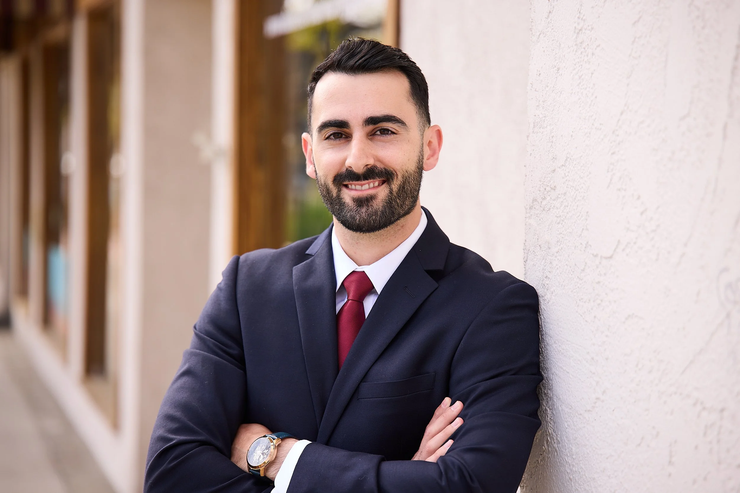 Dr. Arbi Zadoorian, O.D., in a navy suit and red tie, arms folded, leaning against a pale stucco wall outside
