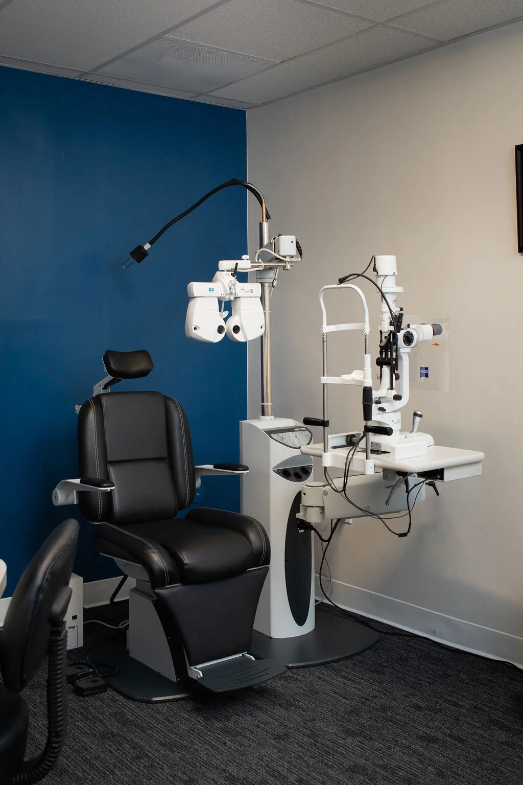 An examination room at New Vision Optometry, a black patient chair and a slit lamp set against the practice's cobalt blue wall