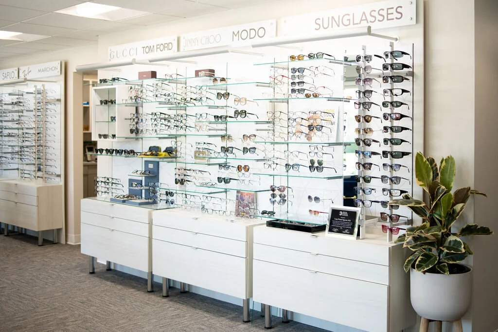 The sunglass wall at New Vision Optometry, glass shelves of sunglasses and eyeglass frames under brand signage, with a rubber plant beside the cabinets