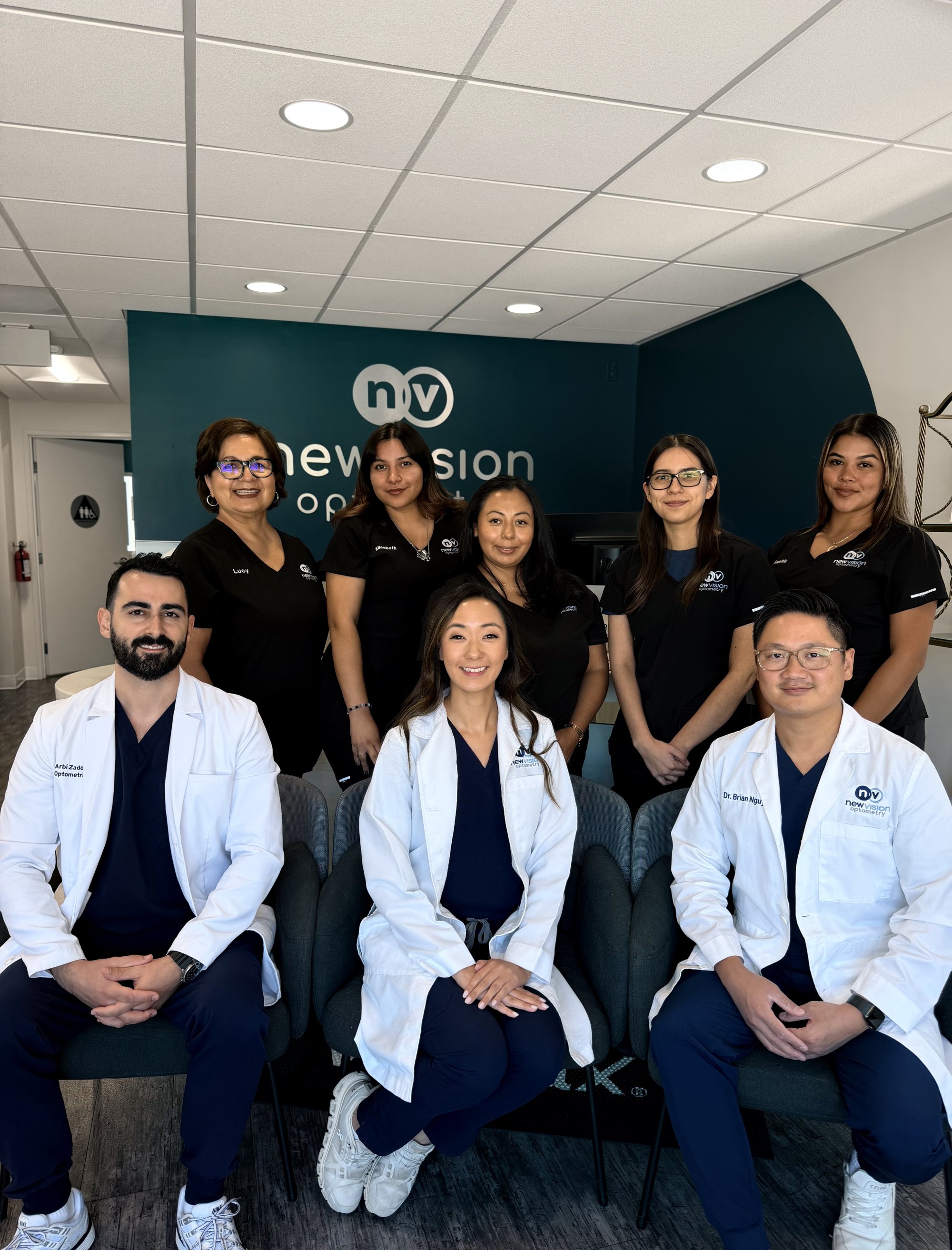 The three New Vision Optometry doctors seated in white coats at the practice mark on the teal wall, the staff standing behind them