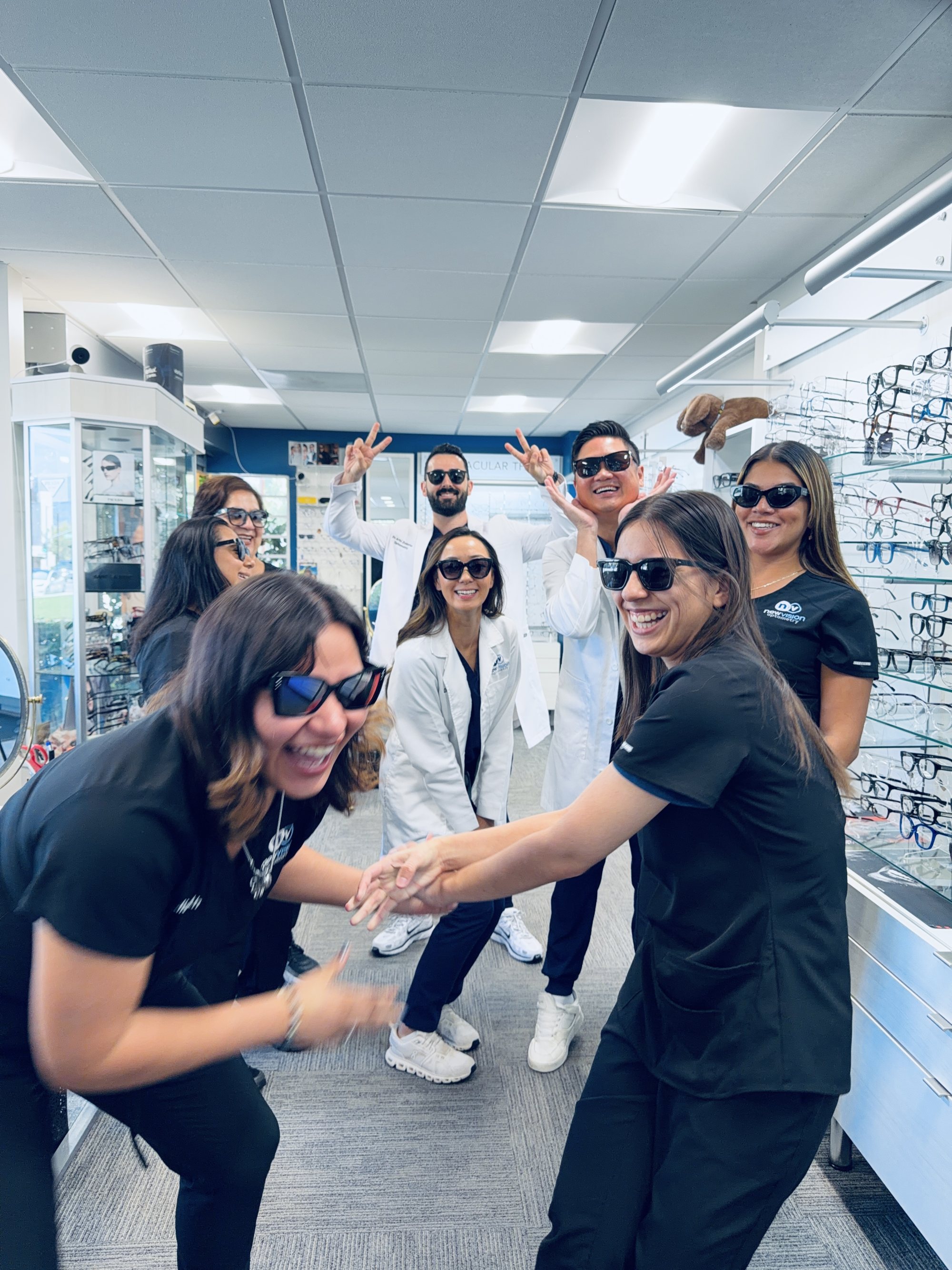 The New Vision Optometry team laughing in the optical in sunglasses from the frame wall, the three doctors and the staff